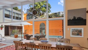 A dining area with a wooden table and chairs, large windows, and potted plants. - Characterful 4BR home w/ pool, full of artwork w/ galleries close by (Paddington)