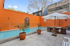 Een buitenzwembad met betegelde vloer, plantenbakken, een eettafel met stoelen en een grote parasol.