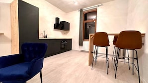 Un soggiorno moderno con una poltrona blu, un'area pranzo con due sgabelli e una cucina con pensili scuri e un televisore nero.