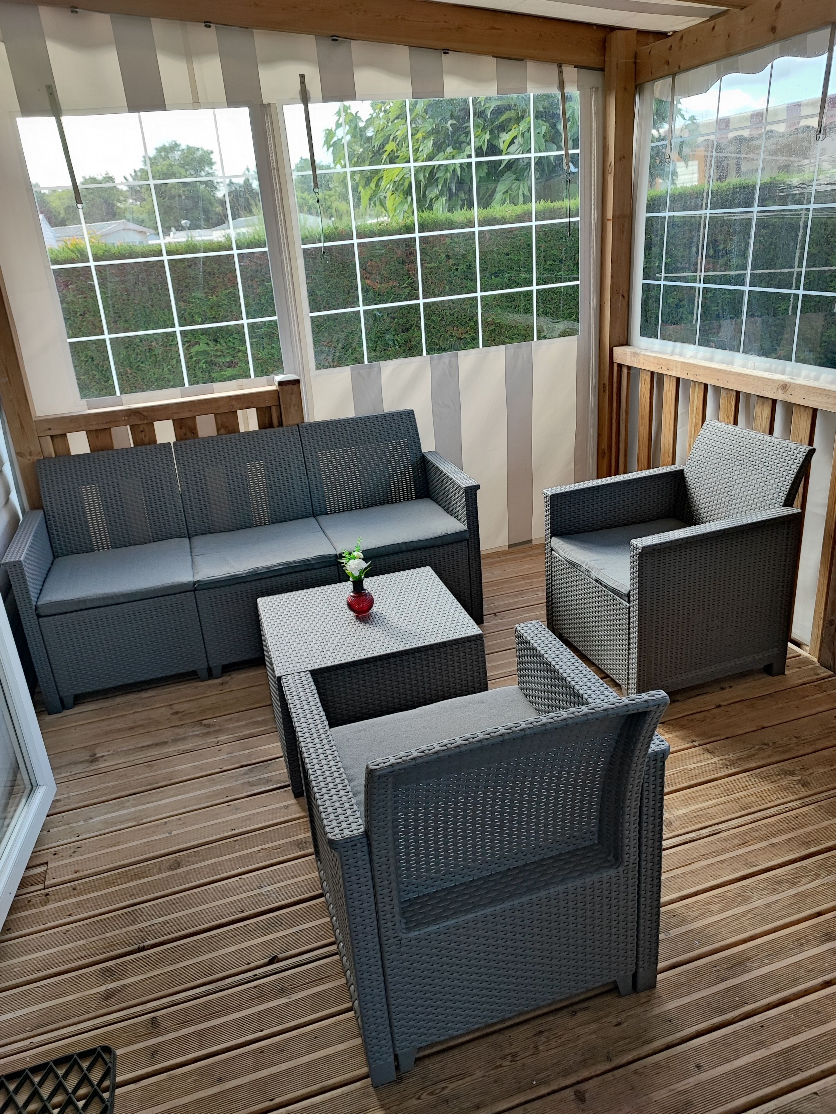 A covered outdoor seating area with a sofa and two chairs, a small table with a vase, and a view of greenery through large windows.