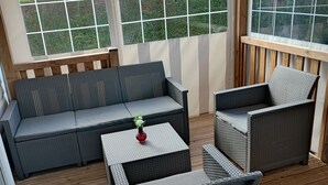 Un espace extérieur aménagé, abrité, avec un canapé et deux fauteuils, une petite table sur laquelle est posé un vase, et une vue sur la verdure grâce à de grandes fenêtres.