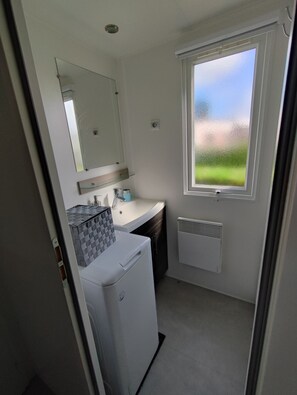 Une petite salle de bain avec un lavabo blanc, un miroir et une fenêtre donnant sur un espace verdoyant.