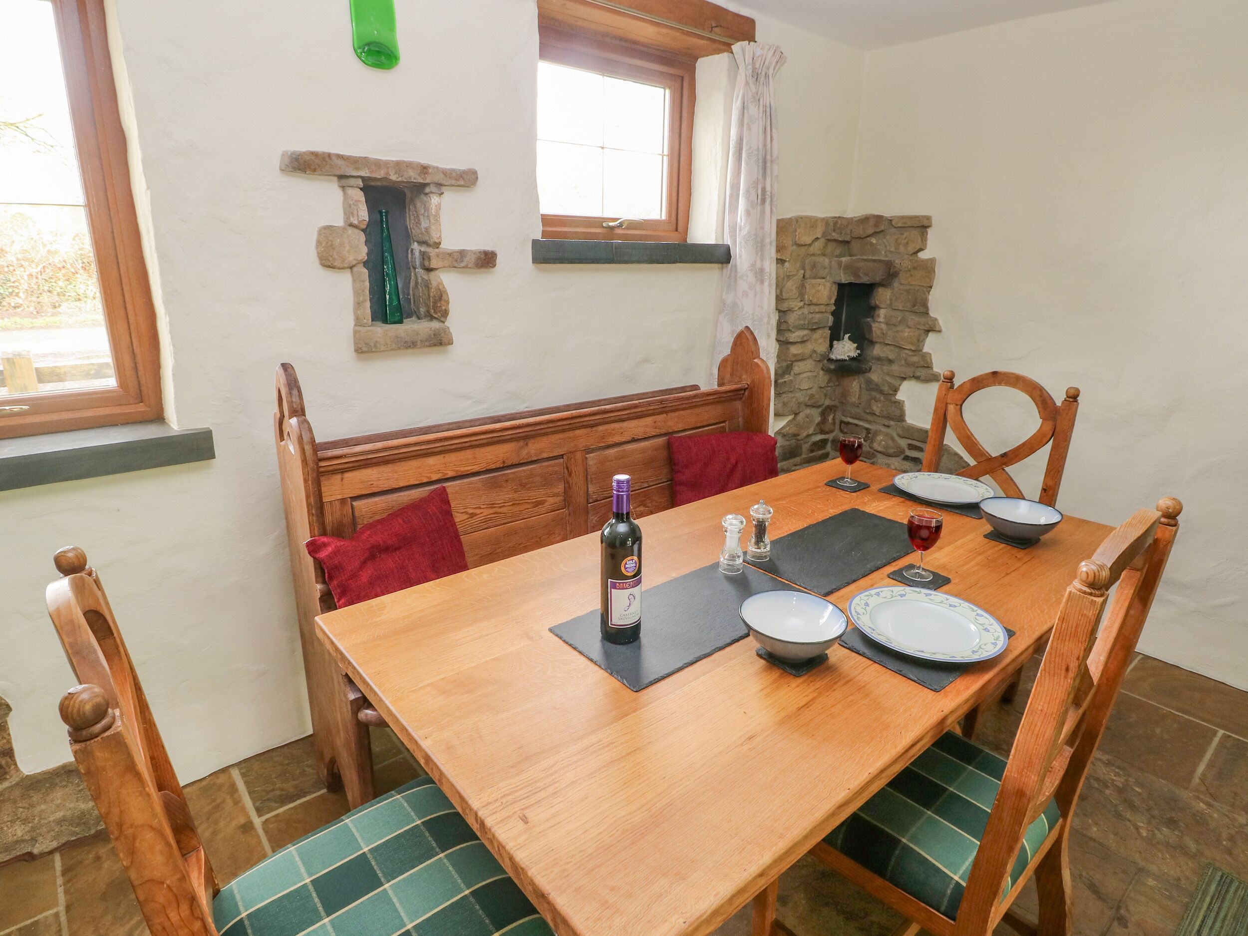Un comedor con una mesa de madera dispuesta para cuatro personas, incluyendo platos, una copa de vino y una botella. Hay dos sillas de madera con cojines verdes. Se aprecia una chimenea de piedra al fondo.