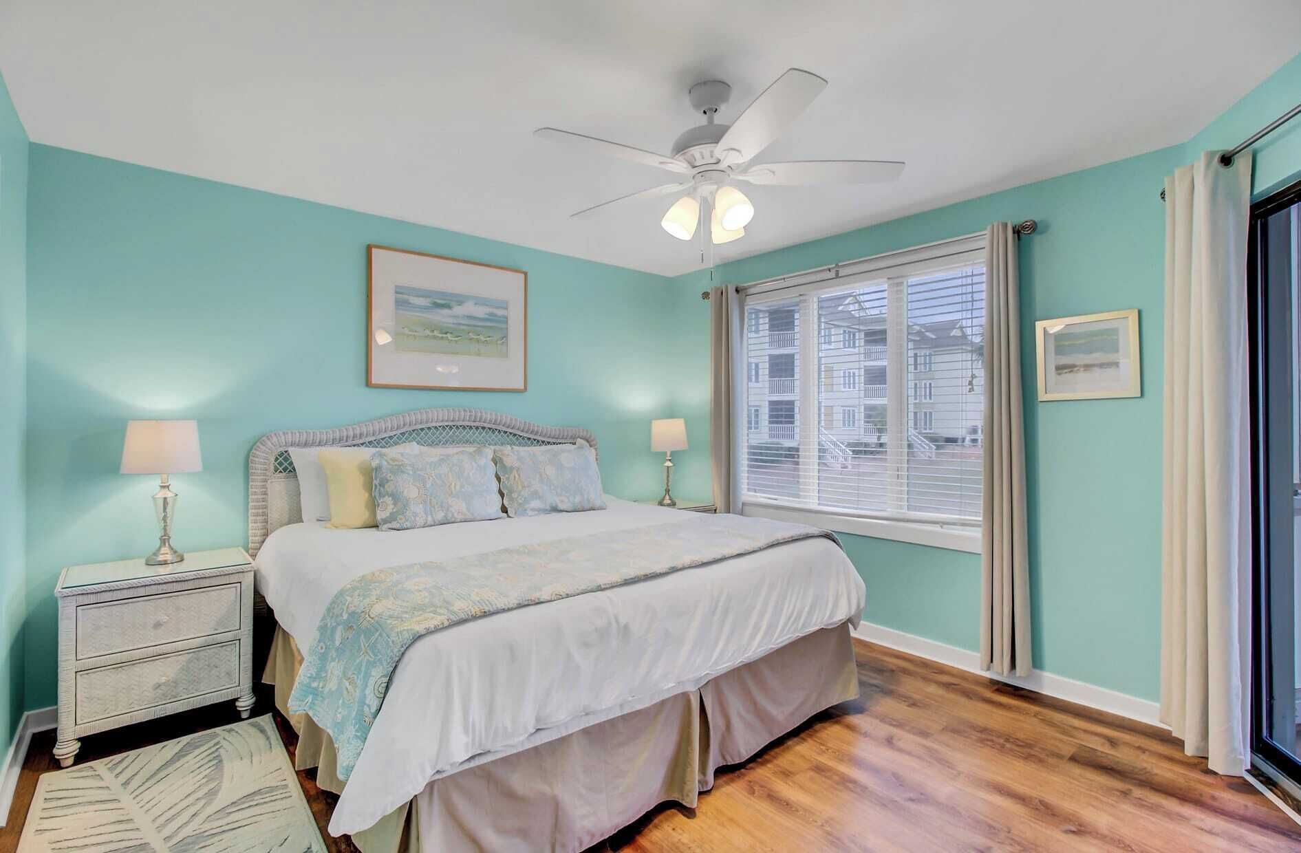 A bedroom with a bed, nightstands, a ceiling fan, a window with blinds, and a view of buildings outside.