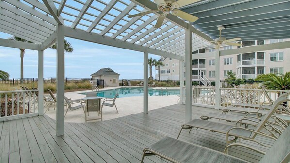 A covered outdoor pool area with lounge chairs and a building in the background.
