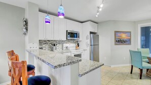 Una cocina moderna con armarios blancos, encimera de granito, microondas, refrigerador y luces colgantes.