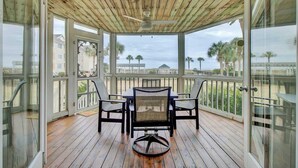 Un espacio al aire libre techado con techo de madera, una mesa con cuatro sillas y vistas a la playa y palmeras.