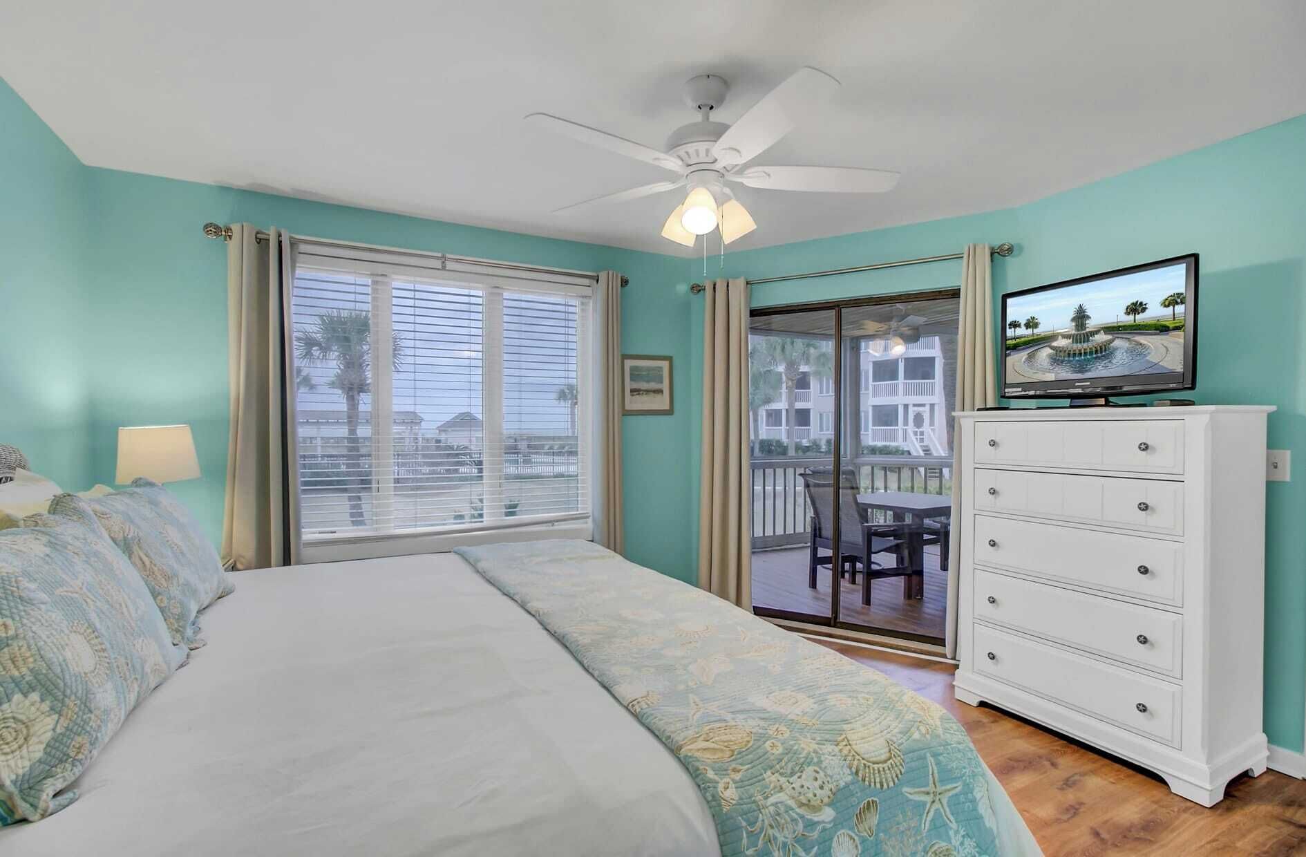 Une chambre avec un lit, un ventilateur de plafond, une télévision, une commode et un balcon donnant sur des immeubles.