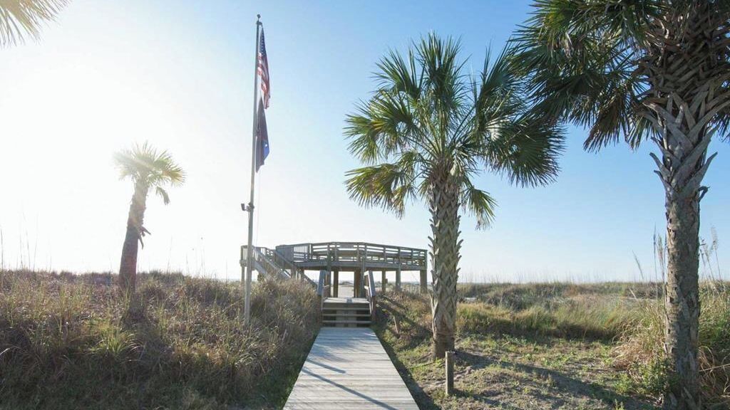 A wooden boardwalk leading to a covered structure with an American flag, surrounded by palm trees.