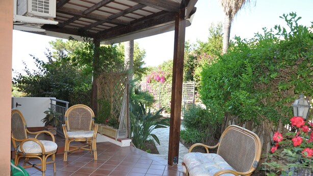 A covered patio with wicker chairs and a table, surrounded by lush greenery and flowering plants.