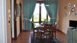 A dining area with a wooden table and chairs, a TV on a side table, and a window with curtains.