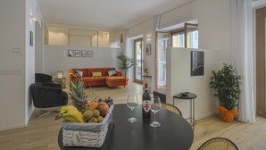 Un vaste espace de vie comprenant une table à manger dressée pour deux personnes, une chaise noire, un panier en osier rempli de fruits, une bouteille de vin et une plante en pot.