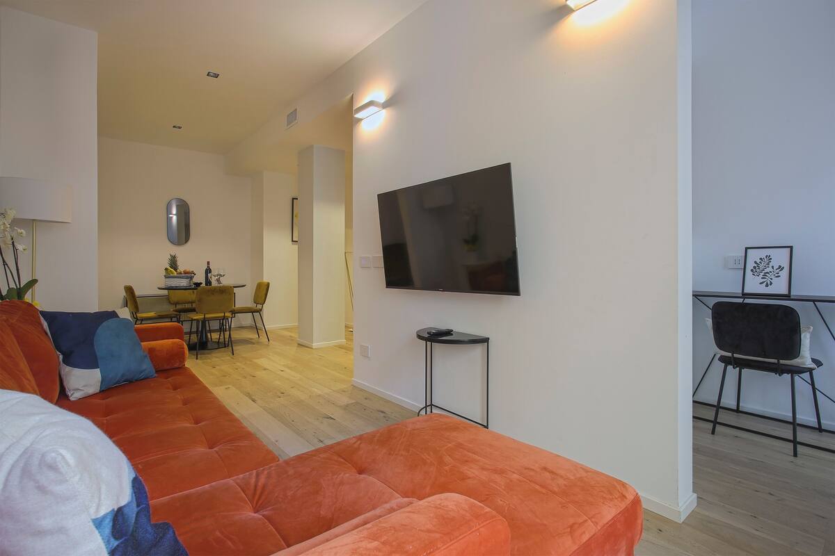 Ein modernes Wohnzimmer mit einem großen, orangefarbenen Sofa, einem flachbildfernseher an der Wand und einem Essbereich mit gelben Stühlen und einem Tisch.