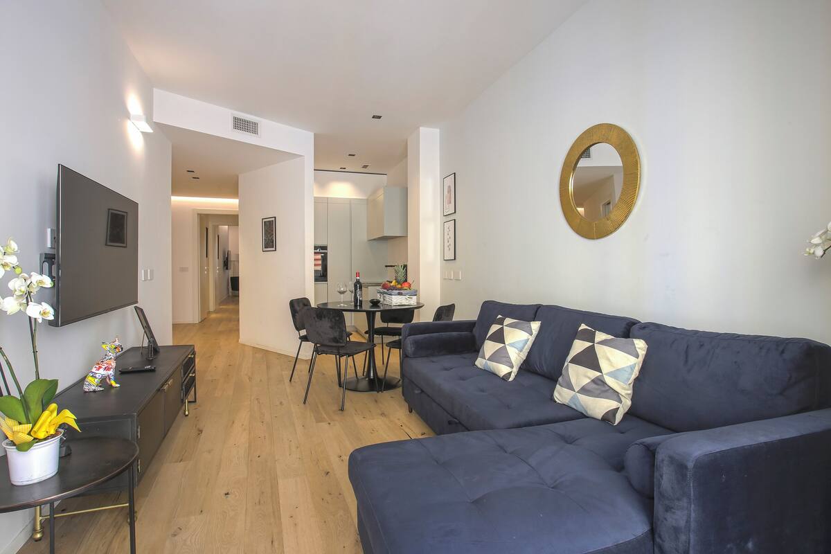 Ein modernes Wohnzimmer mit einem dunkelblauen Sofa, einem an der Wand montierten Flachbildfernseher und einem Essbereich mit Tisch und Stühlen.