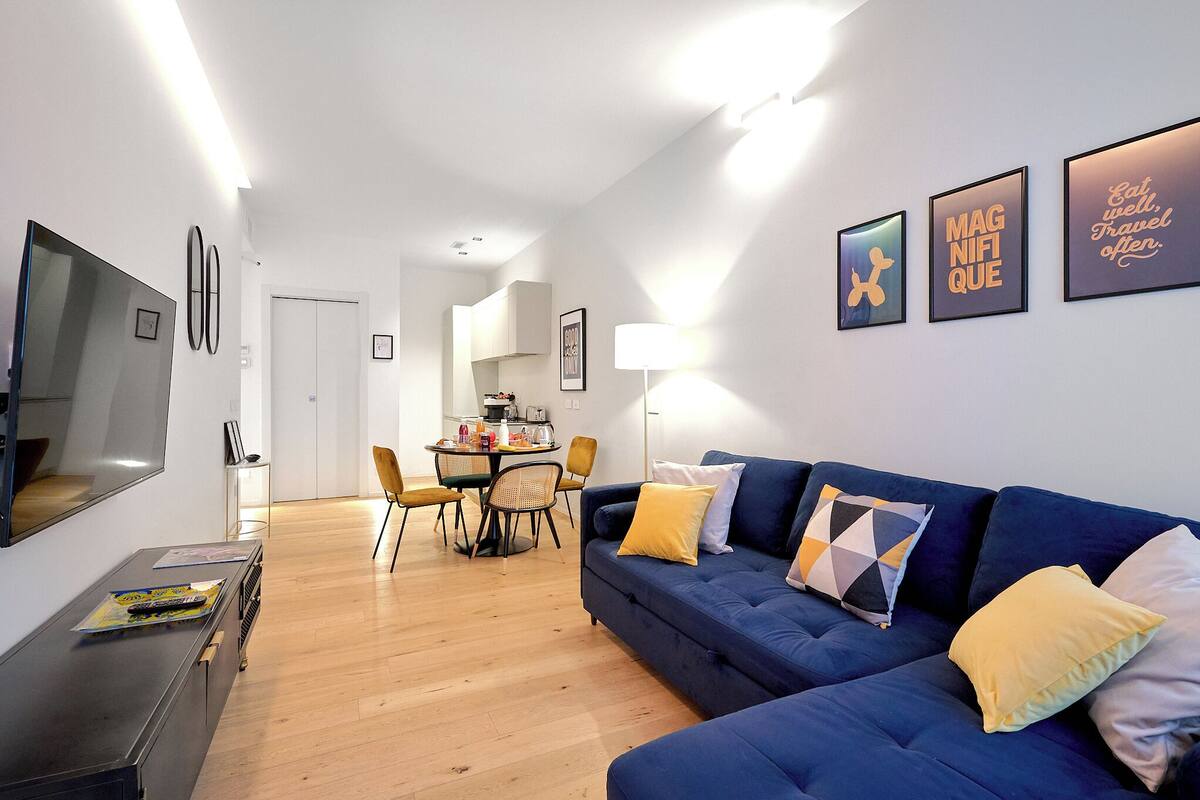 Ein modernes Wohnzimmer mit einem blauen Sofa, gelben Kissen, einem Flachbildfernseher und einem Essbereich mit Tisch und Stühlen.