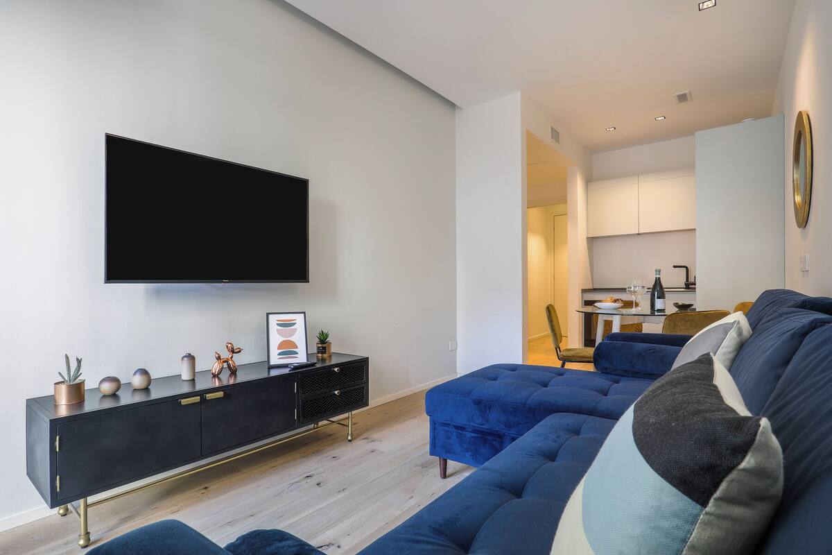 Ein modernes Wohnzimmer mit einem großen Flachbildfernseher an der Wand, einem schwarzen Konsolentisch mit Dekoration, einem blauen Sofa mit gemusterten Kissen und einem Blick auf eine Küchenzeile im Hintergrund.
