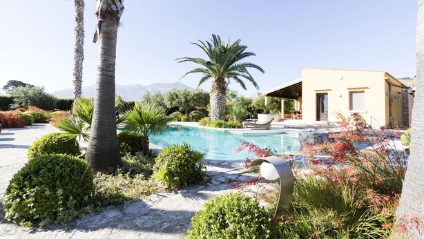 Ein Poolbereich mit freiem Blick auf den Pool, umgeben von Pflanzen und einem Gebäude mit überdachter Terrasse.