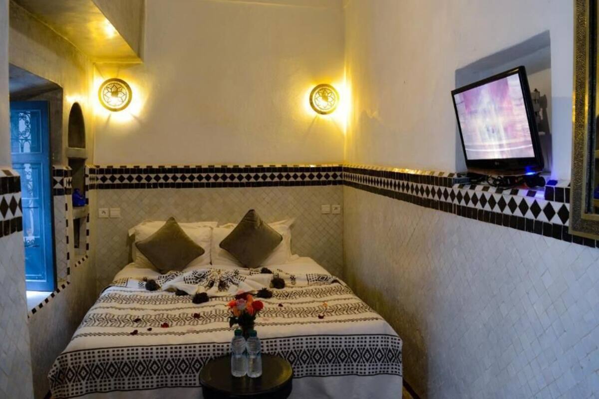 A hotel room with a bed, two wall-mounted lights, a TV on a stand, and a small table with two bottles.