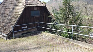 A wooden cabin with a metal staircase leading up to a window, surrounded by trees and a mountainous backdrop.