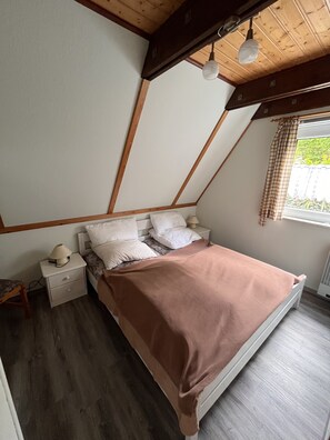 Relax in this serene bedroom with a sloped ceiling, featuring a comfortable bed and a window offering a view of lush greenery.