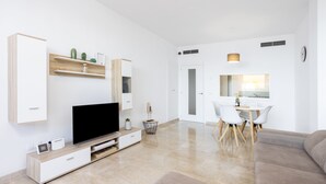 Un soggiorno moderno con una TV a schermo piatto, mobili in legno, un'area pranzo con tavolo e sedie bianche e una cucina visibile attraverso una porta aperta.