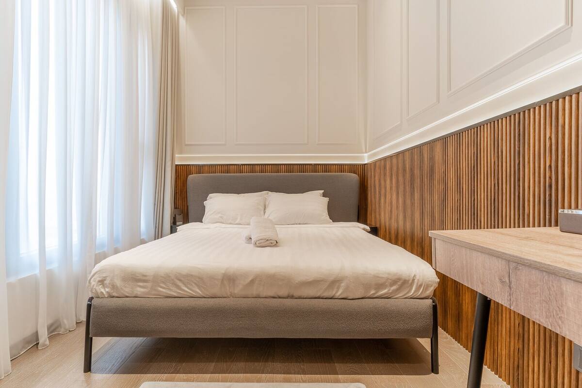 A neatly made bed with a grey headboard, a wooden panel wall, and a wooden floor.