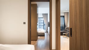 Una habitación de hotel moderna con una puerta de madera, una cama con ropa blanca, una mesita y vistas a la ciudad a través de amplios ventanales.