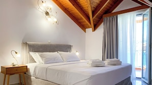 A bedroom with a bed, bedside lamps, a nightstand, and a view of the outside through a window. - Rouchotas Apartments (Kefalonia)