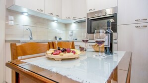 A modern kitchen with white cabinets, a stainless steel oven, and a dining table with a glass top. - Rouchotas Apartments (Kefalonia)