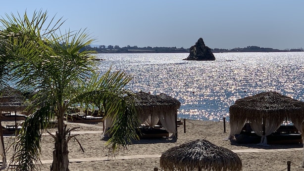 A beach with thatched umbrellas and palm trees, a rocky outcrop in the water, and a clear sky. - Rouchotas Apartments (Kefalonia)