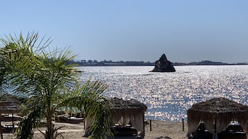 Uma praia com guarda-sóis de palha e palmeiras, um afloramento rochoso na água e um céu claro.