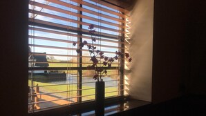 A window with horizontal blinds, a vase with flowers, and a view of a grassy area and a building in the distance.
