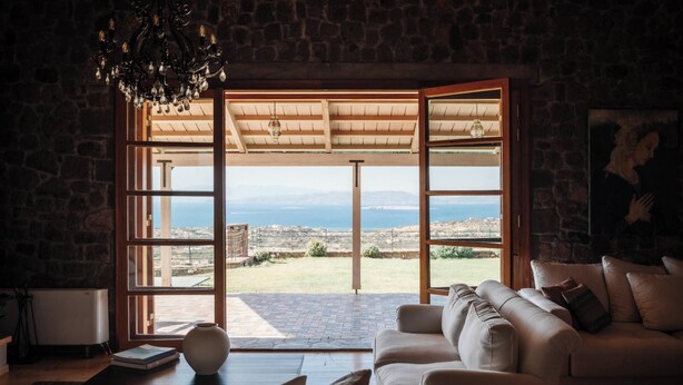 A room with a stone wall, a white radiator, a white sofa, and a view of the sea through large glass doors. - Terra Cassa Private Villa (Aegina)