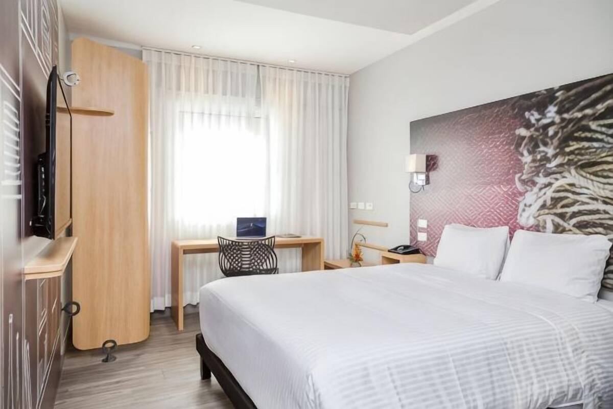 Ein Hotelzimmer mit Bett, Schreibtisch, Stuhl und einem großen Fenster mit Vorhängen.