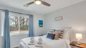 Une chambre à coucher comprenant un lit, une table de chevet, une lampe, une fenêtre avec des rideaux et un ventilateur de plafond.