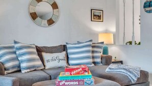 Un salon avec un canapé gris, des coussins à rayures, un miroir rond et une pile de jeux de société.