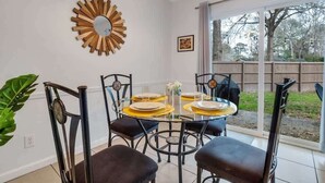 Un coin repas avec une table ronde dressée pour quatre personnes, entourée de chaises. Un miroir décoratif est accroché au mur, et une pancarte portant une citation inspirante est visible.