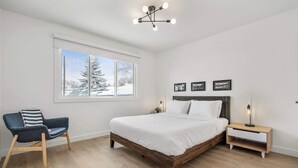 A bedroom with a large bed, a chair, a nightstand, and a window with a view of snow-covered trees. - Welcome to this charming, newly renovated home in the heart of Bowness, one of Calgary’s most beloved and character-filled neighbourhoods. Known for its small-town feel, access to nature, and vibrant local scene, Bowness is the perfect home base. (Calgary)
