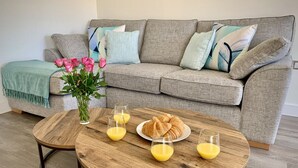 A living room with a grey sofa, two round wooden coffee tables, a vase with pink flowers, and croissants on a plate. - Tolcarne - Perranporth Cornwall (Perranporth)