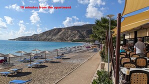 A beachside restaurant with outdoor seating and umbrellas, overlooking a beach with clear blue water and a mountainous backdrop. - Pissouri stylish and spacious contemporary 2 bedroom apartment (Pissoúri)