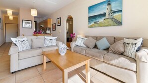 A living room with two sofas, a coffee table, a painting of a lighthouse, and a kitchen area in the background. - Pissouri stylish and spacious contemporary 2 bedroom apartment (Pissoúri)