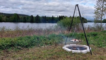 En lägereld med ett stativ, omgiven av gräs och buskar, nära en sjö med träd i bakgrunden.