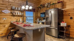 Una cocina con paredes de madera, un refrigerador de acero inoxidable, una encimera de granito, taburetes de bar y un letrero que dice 'KITCHEN'S FAMILIES TOGETHER'.
