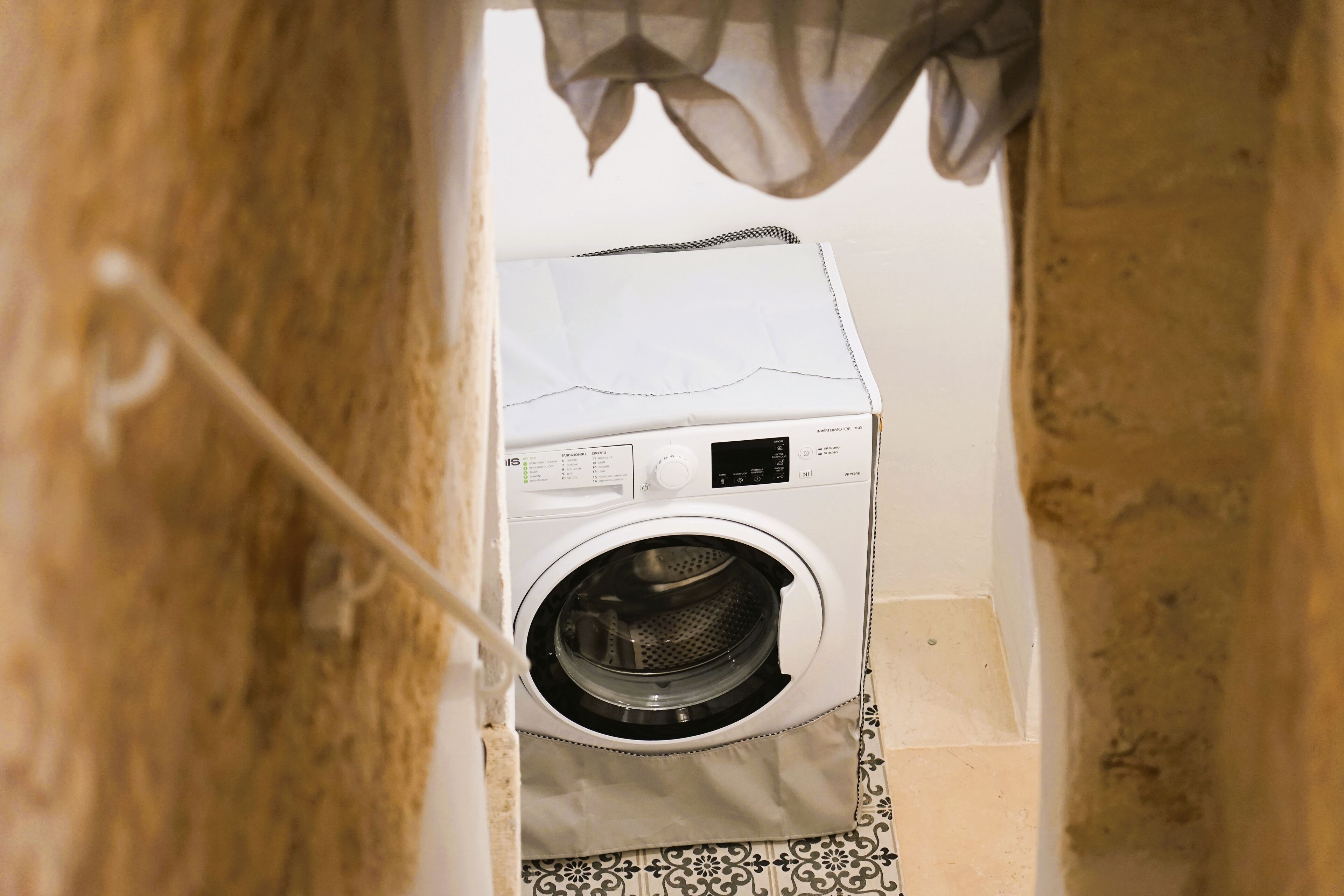 Una lavadora blanca de carga frontal, compacta, en un cuarto de lavado estrecho con pisos de baldosas estampadas.