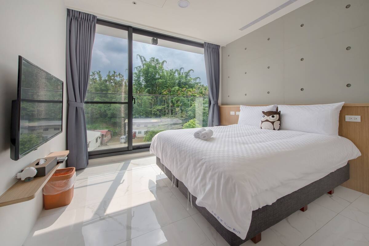 A modern hotel room with a large bed, a flat-screen TV, a balcony with a view of greenery, and a wooden headboard.
