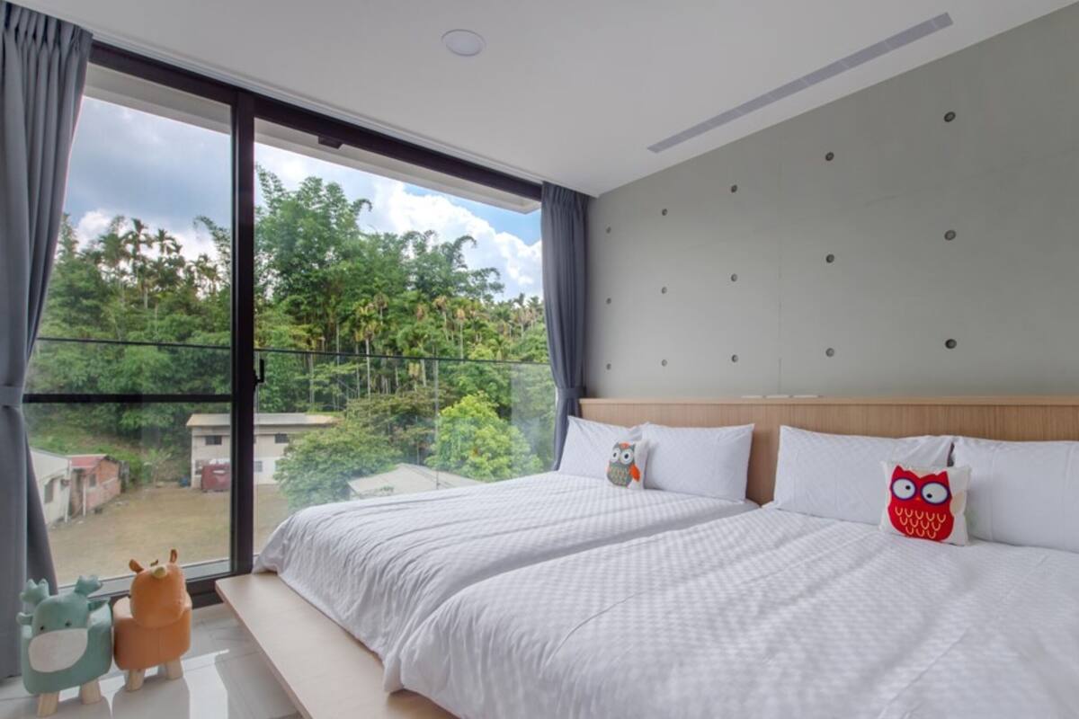 A modern hotel room with a large window, a bed with white bedding, and a view of greenery outside.