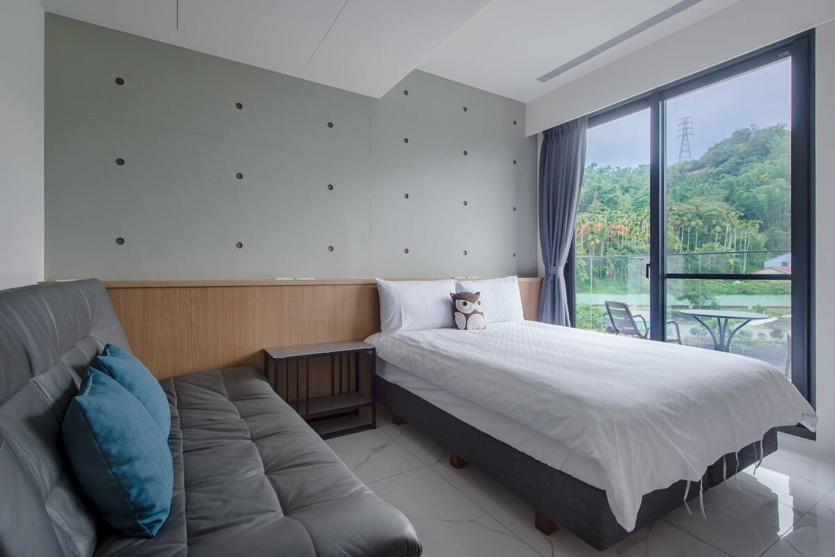 A modern hotel room with a bed, a sofa, a small table, and a large window offering a view of greenery.