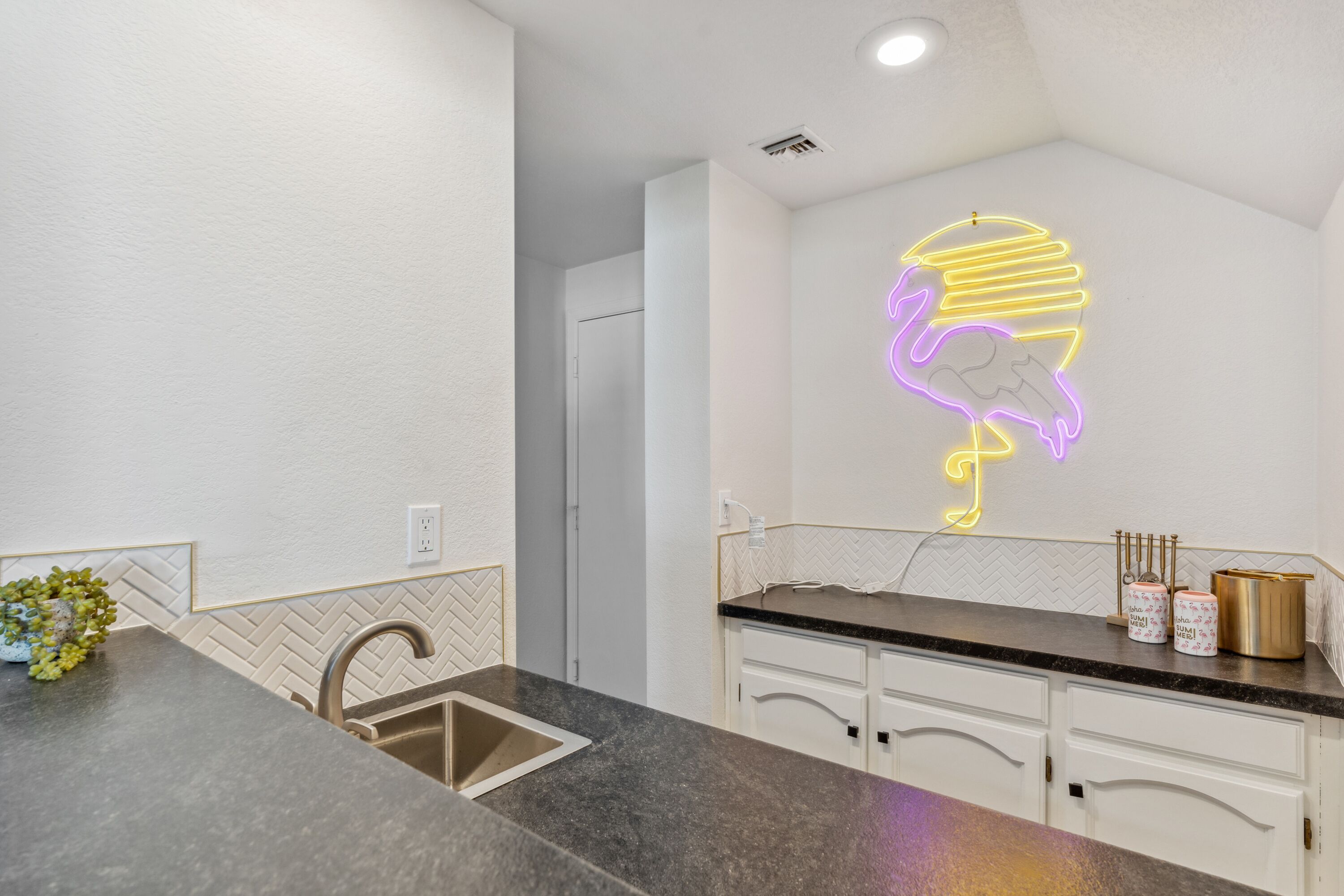 A modern kitchen with a herringbone-patterned backsplash, a stainless steel sink, and a neon light shaped like a flamingo.