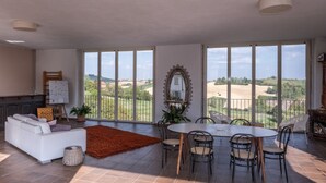 Un vaste espace de vie comprenant une table à manger et des chaises, un canapé blanc et une grande fenêtre donnant sur la campagne.