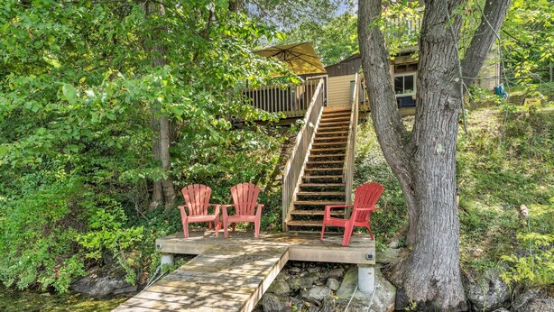 A wooden deck with a staircase leading to a deck chair area surrounded by trees. - Black Rapids Retreat | Waterfront Cozy Cottage (Leeds and the Thousand Islands)
